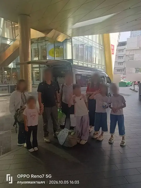 2026년 10월 5일 오후 4시 3분, 방콕에서 금색 기둥이 있는 건물 입구 캐노피 아래, 뒷문을 연 밴 옆에 여러 아이와 짐과 함께 모여 선 가족