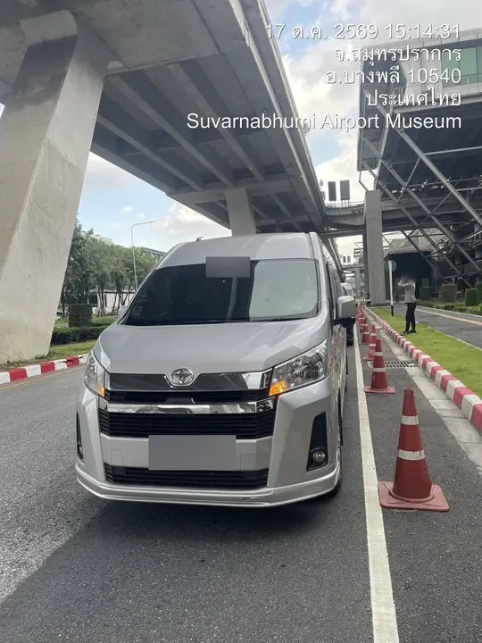 The new-model silver Toyota Commuter stands head-on in the pickup lane under the elevated roadway at Suvarnabhumi Airport, traffic cones beside it, in Bang Phli, Samut Prakan, at 3:14 p.m. on 17 October 2026.