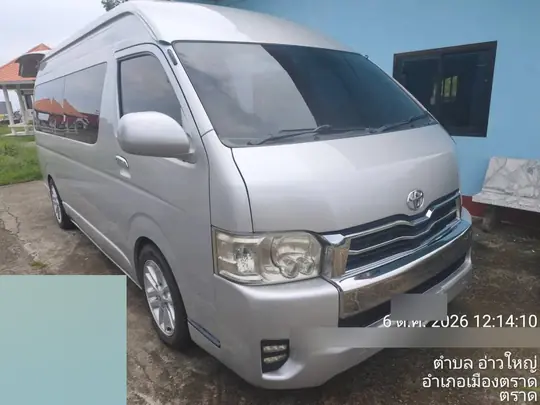 2026년 10월 6일 낮 12시 14분, 뜨랏 므앙뜨랏 아오야이에서 파란 벽과 기와지붕 건물을 배경으로 잔디 위에 정면으로 서 있는 실버 토요타 커뮤터
