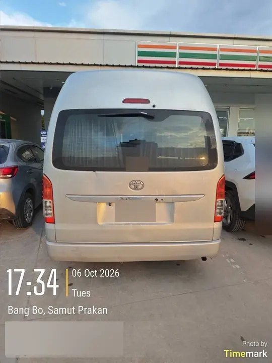 The silver high-roof Toyota Commuter is parked under a convenience-store canopy, seen from the rear with cars on either side, in Bang Bo, Samut Prakan, at 5:34 p.m. on 6 October 2026.