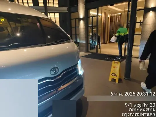 The silver van waits head-on at a hotel entrance at night, the lit glass lobby and a wet-floor sign behind it, on Sukhumvit 20, Khlong Toei, Bangkok, at 8:31 p.m. on 6 October 2026.